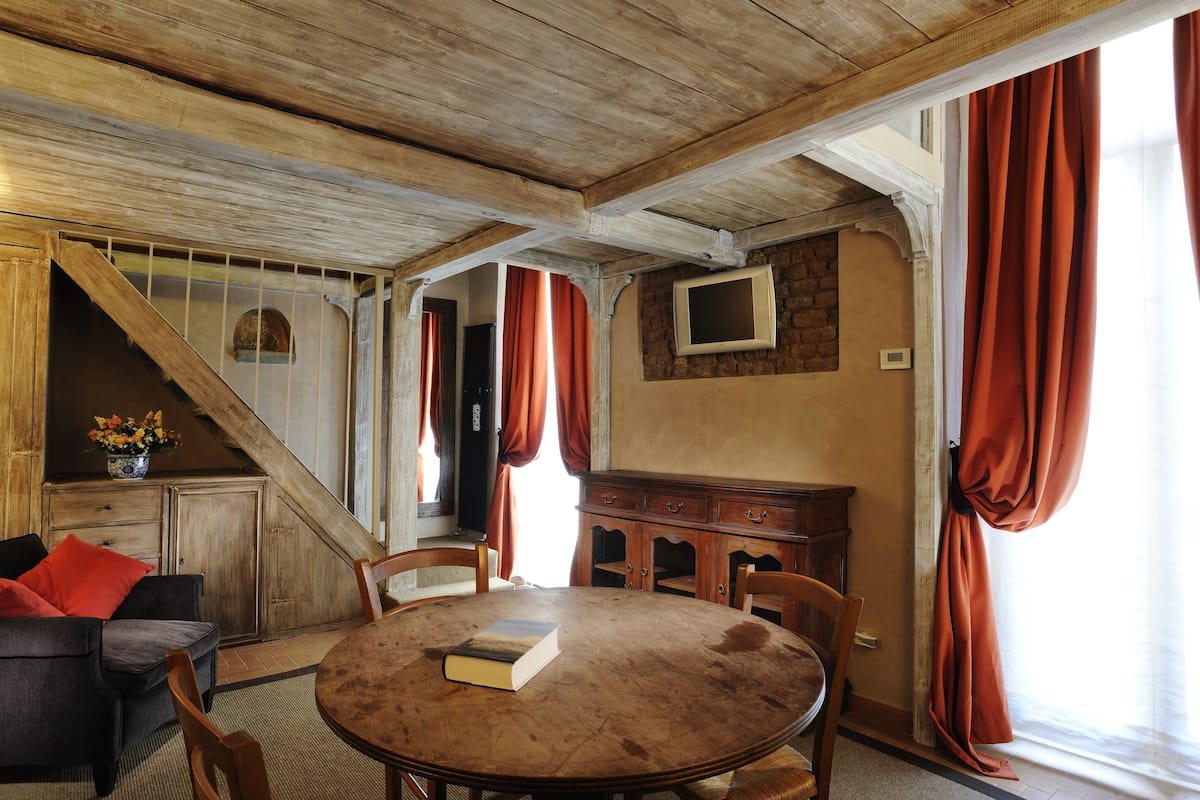 A room with a wooden ceiling, a round wooden table with a book, a sofa, and a TV on the wall.