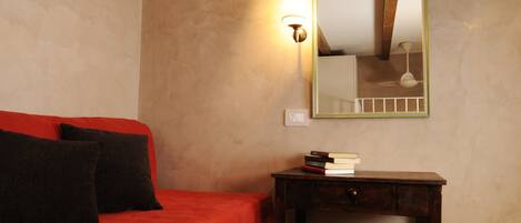 A bedroom with a red bed, wooden beams, a small table with books, and a wall-mounted mirror.
