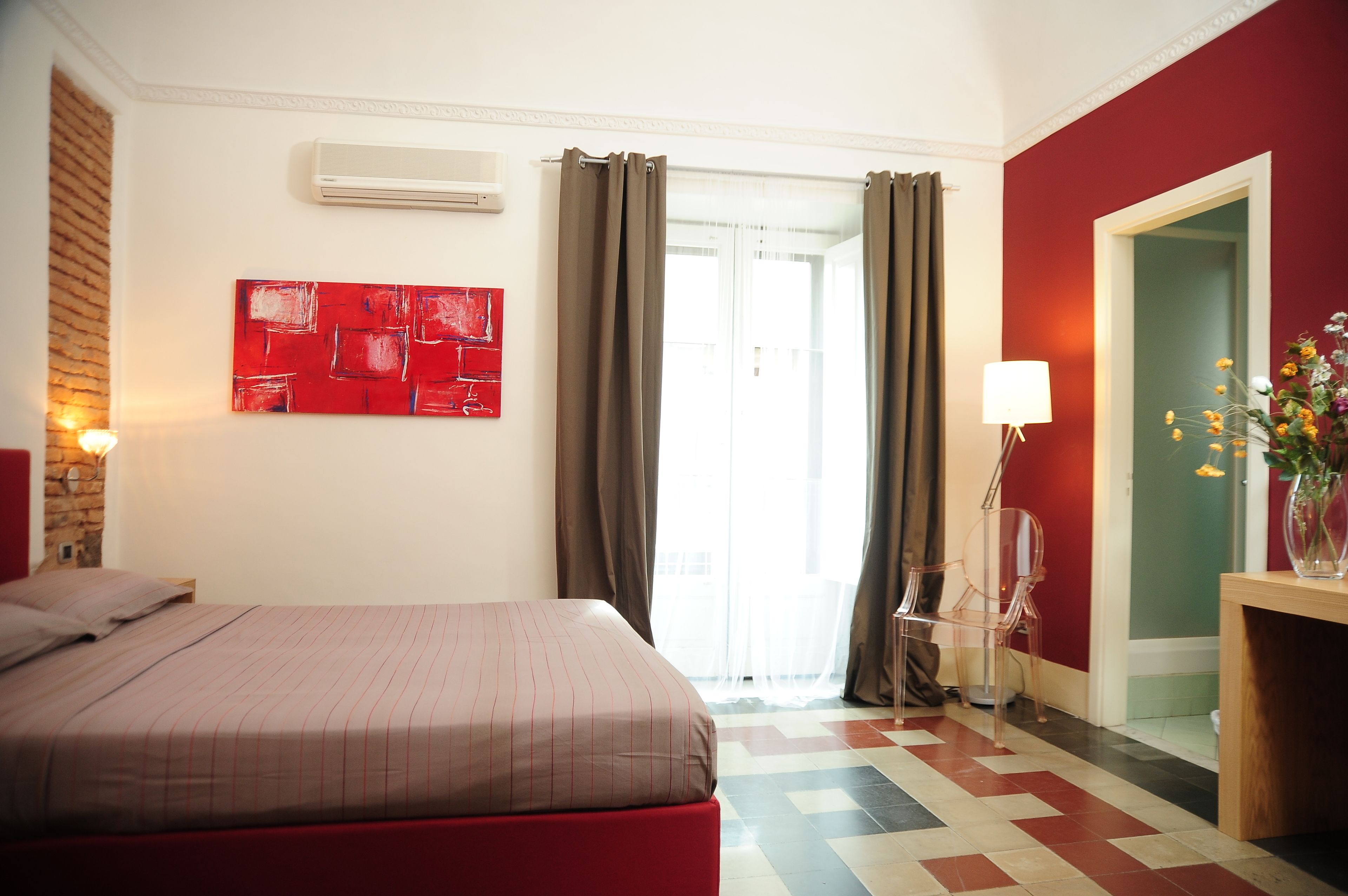 Una habitación de hotel con cama, pared roja, ventana con cortinas, un jarrón con flores y una lámpara.