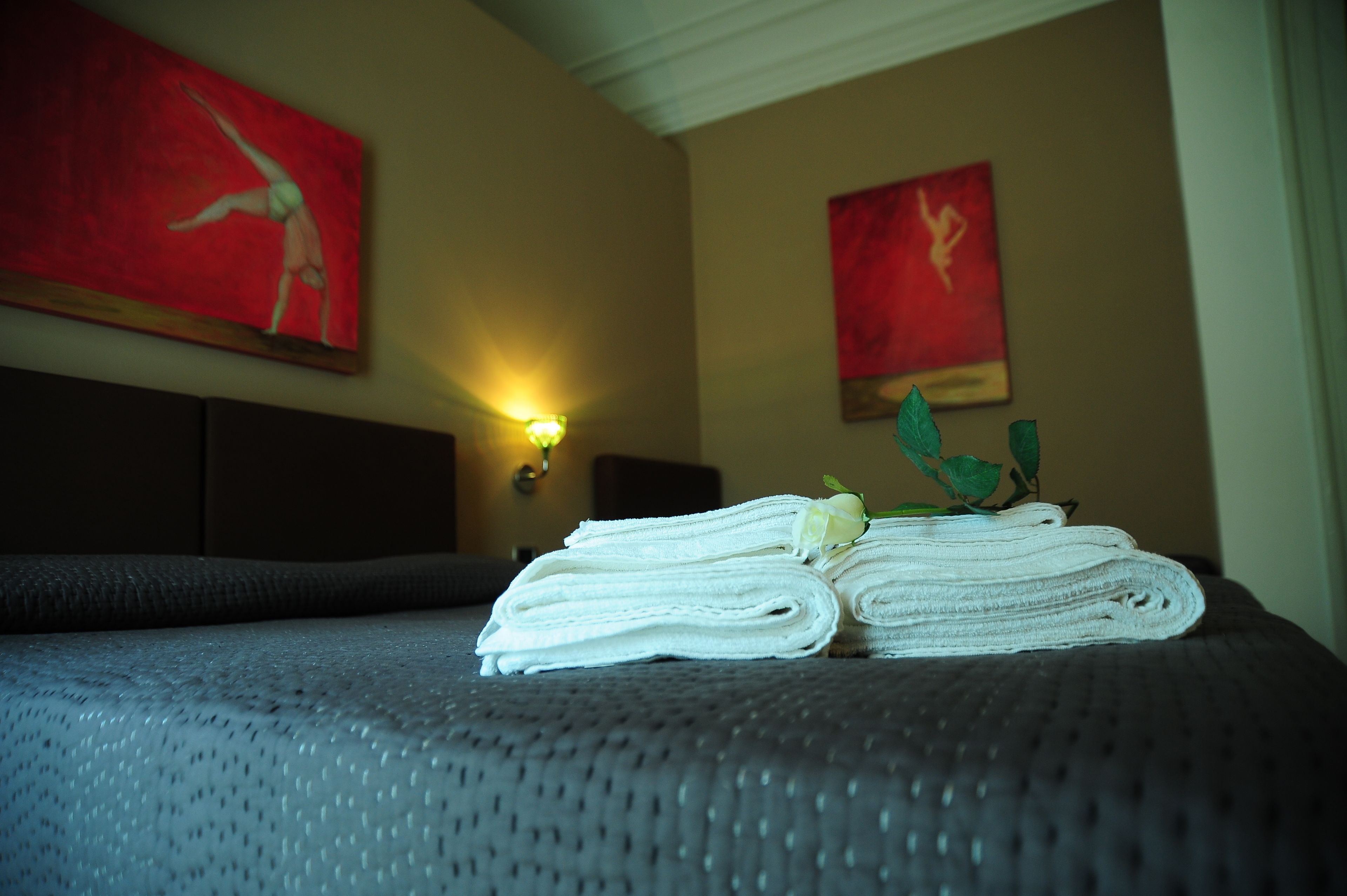 A neatly made bed with folded towels and a rose, a wall-mounted light, and two red abstract paintings.