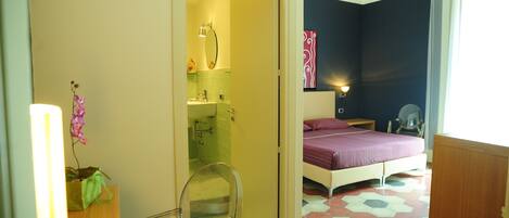 A room with a bed, a mirror, a small table, and a chair. There is a bathroom visible through an open door.