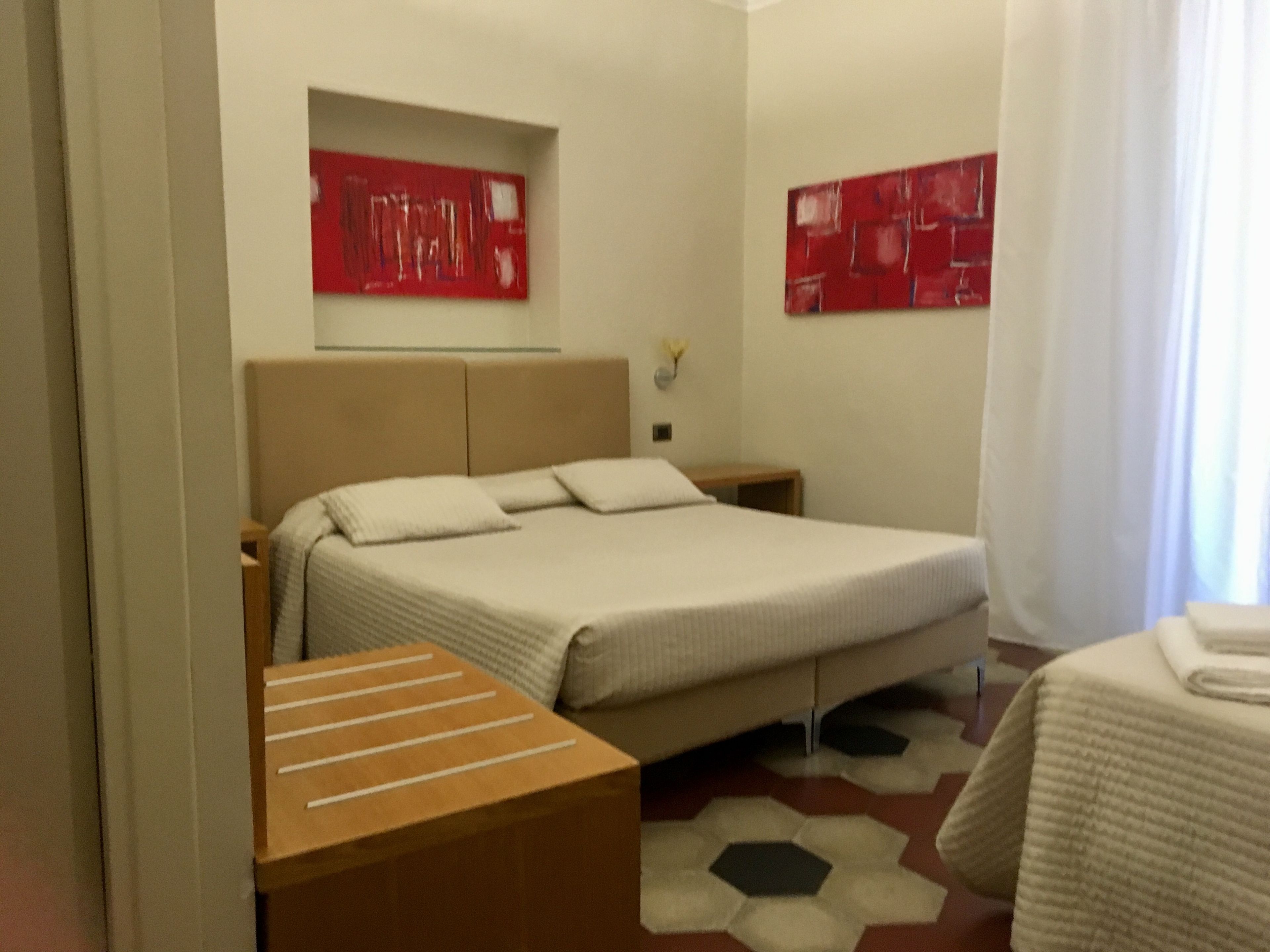 A hotel room with a bed, a nightstand, a wooden side table, and two red abstract paintings on the walls.