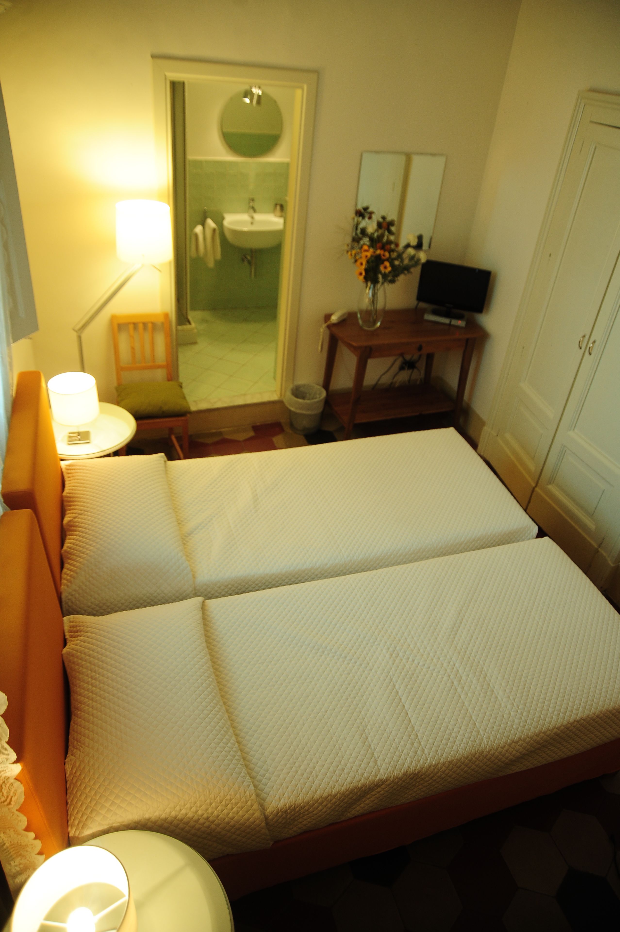 A hotel room with a bed, a bedside lamp, a chair, a small table with a lamp, a mirror, and a bathroom visible through an open door.