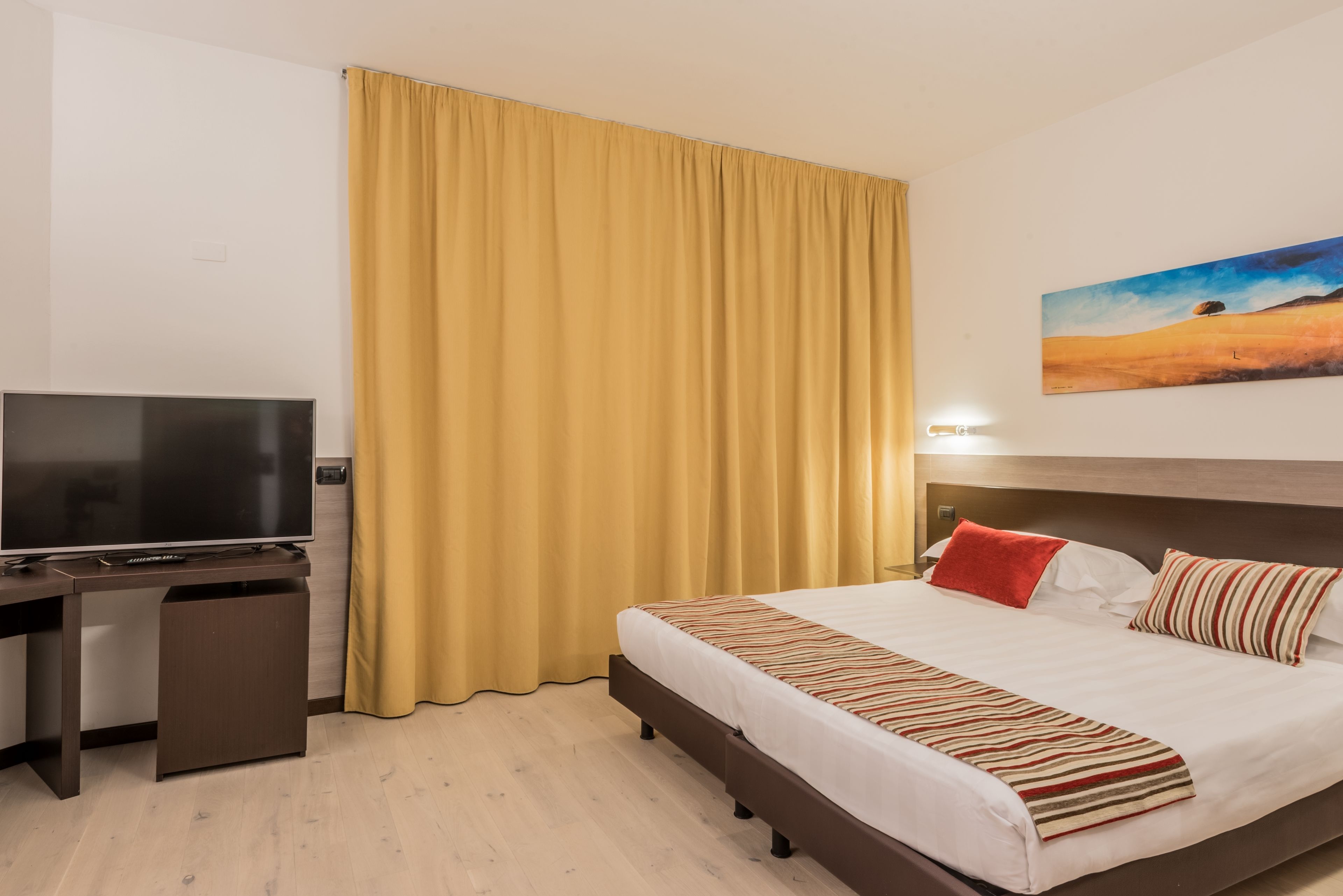 A hotel room with a bed, a TV, a desk, and a painting on the wall.