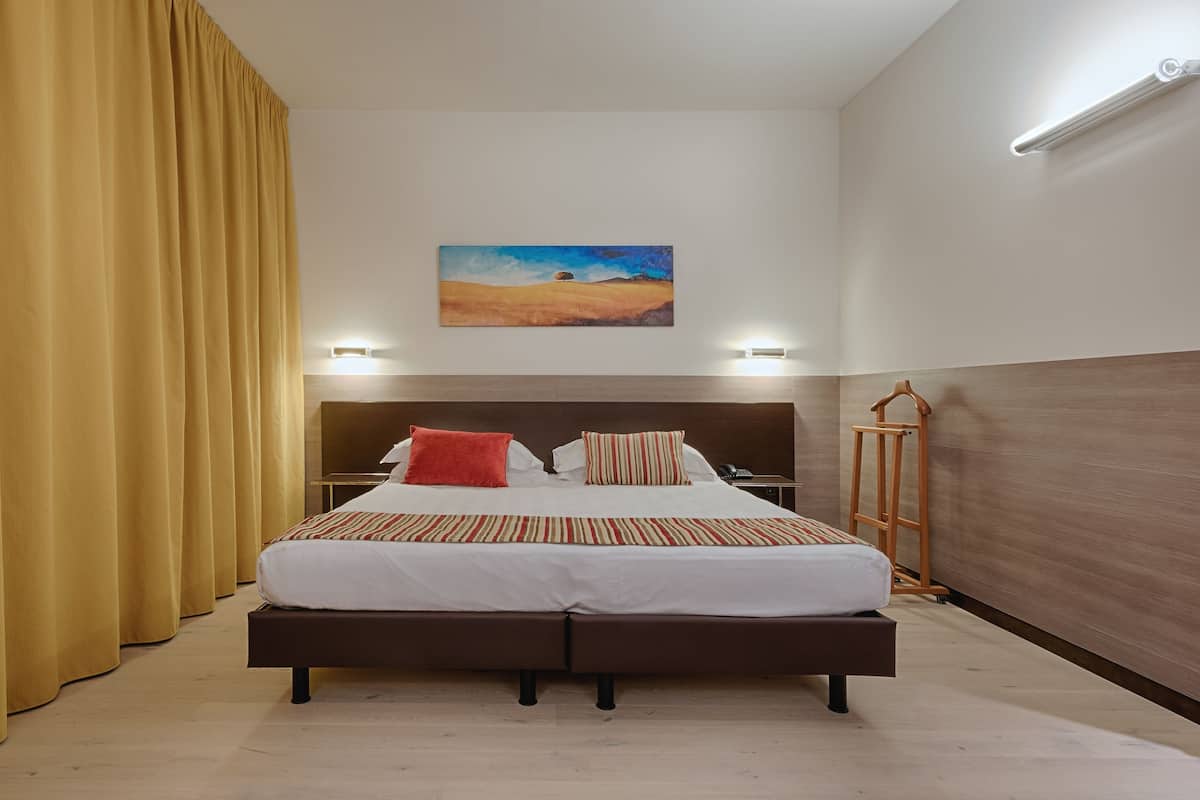 A hotel room with a bed, curtains, a painting, bedside lamps, and a wooden headboard.