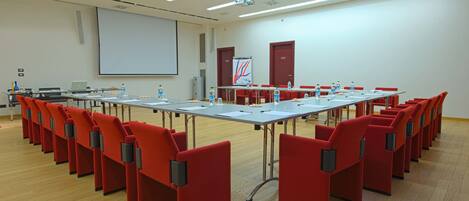 A conference room with rows of red chairs, a long table, a projector screen, and a whiteboard.