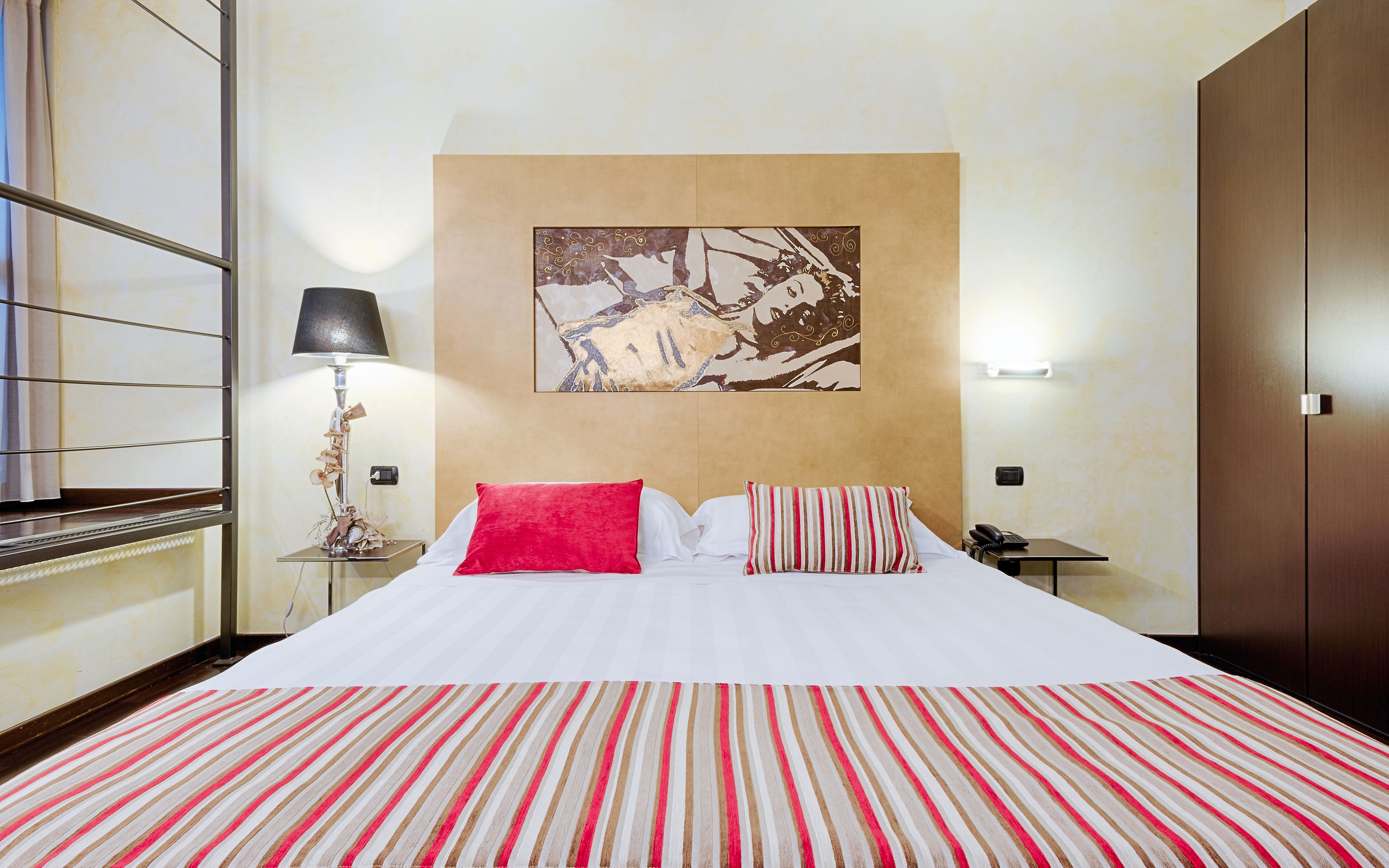A hotel room with a large bed, a nightstand with a lamp, a painting on the wall, and a wardrobe.