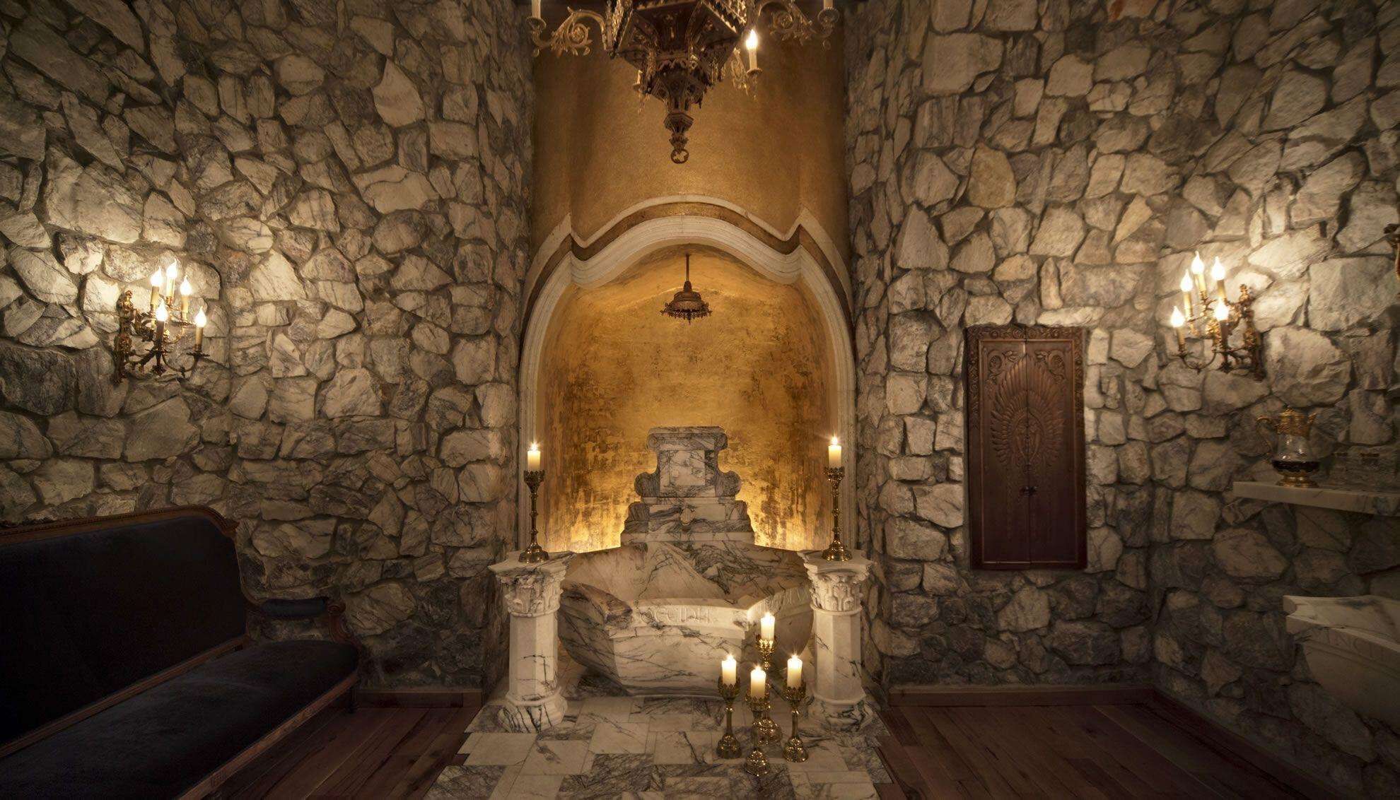 Una habitación de paredes de piedra con un nicho central, una araña y apliques en la pared.