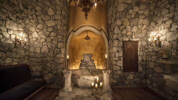 Una habitación de paredes de piedra con un nicho central, una araña y apliques en la pared.