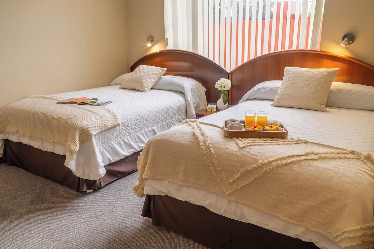 Habitación de hotel con dos camas, cada una con cabecera de madera, ropa de cama blanca y una bandeja con jugo de naranja y fruta sobre una de las camas.