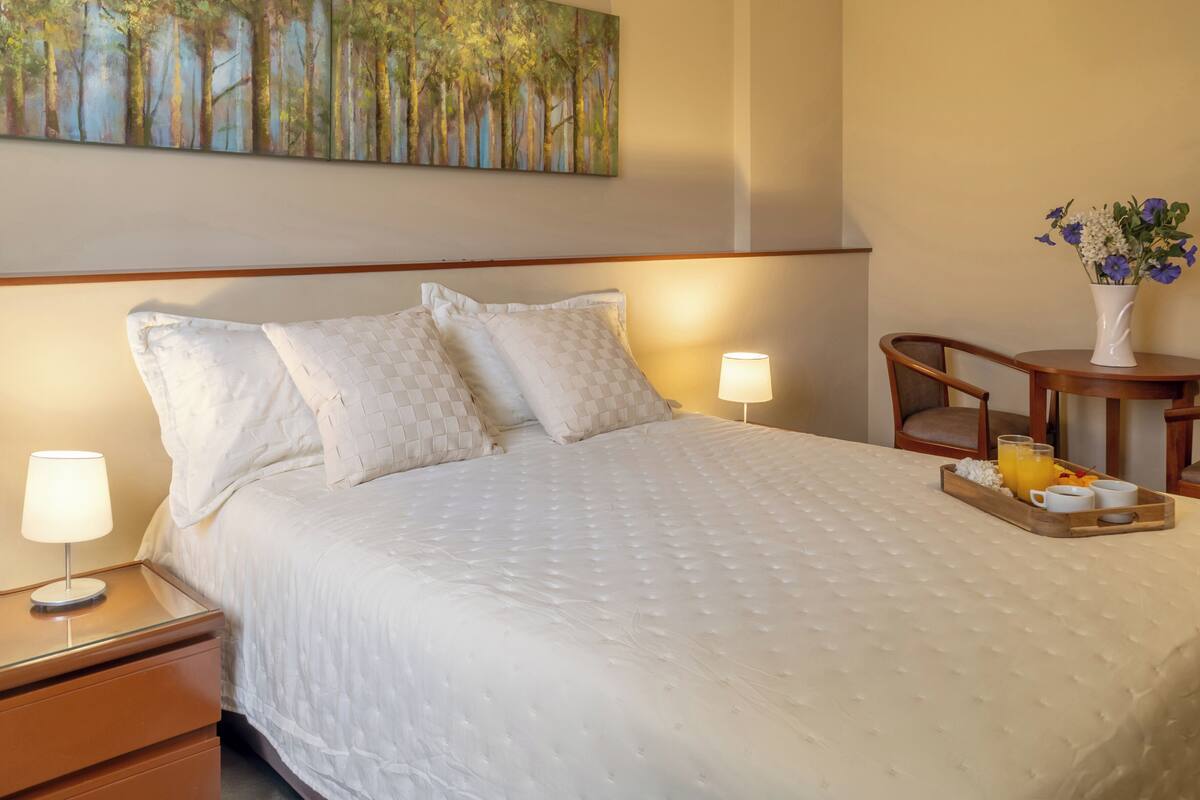 Una cama bien hecha con colcha blanca, una mesita de noche con lámpara, una bandeja con artículos de desayuno y un cuadro de un bosque en la pared.