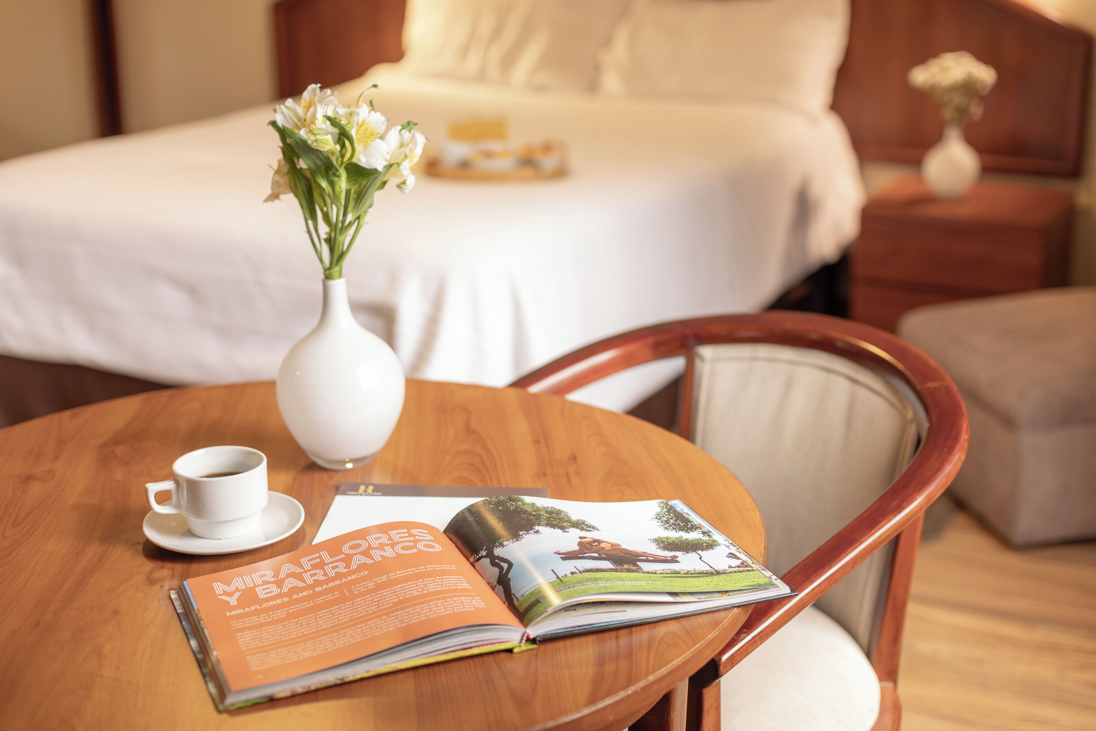Ein Hotelzimmer mit einem Bett, einem Nachttisch mit Blumenvase, einer Kaffeetasse und einer Zeitschrift auf einem Holztisch.