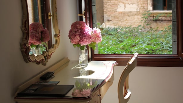 A room with a desk, a vase with flowers, a mirror, and a window with a view of greenery and a building. - Bed & Breakfast Barababao (Venice)