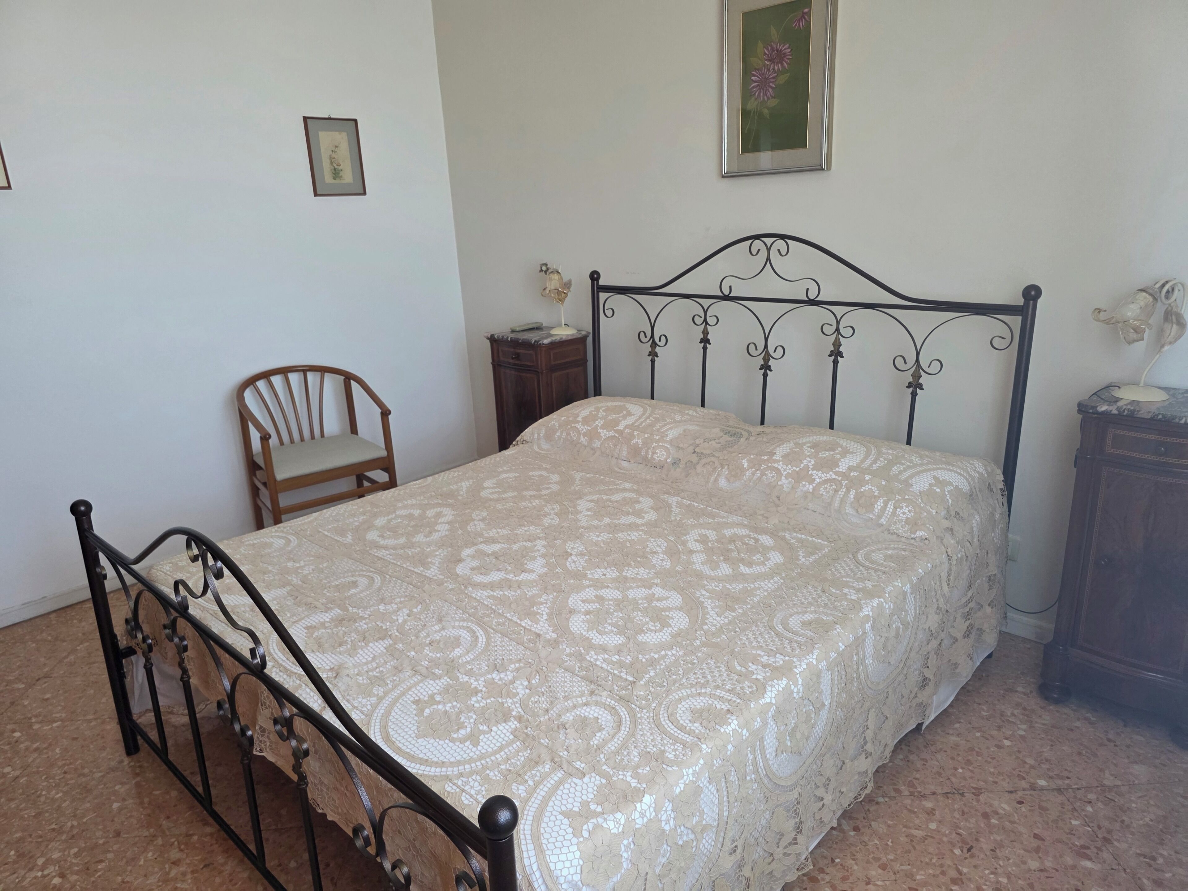A bedroom with a bed, a chair, a nightstand, and a wall-mounted picture.