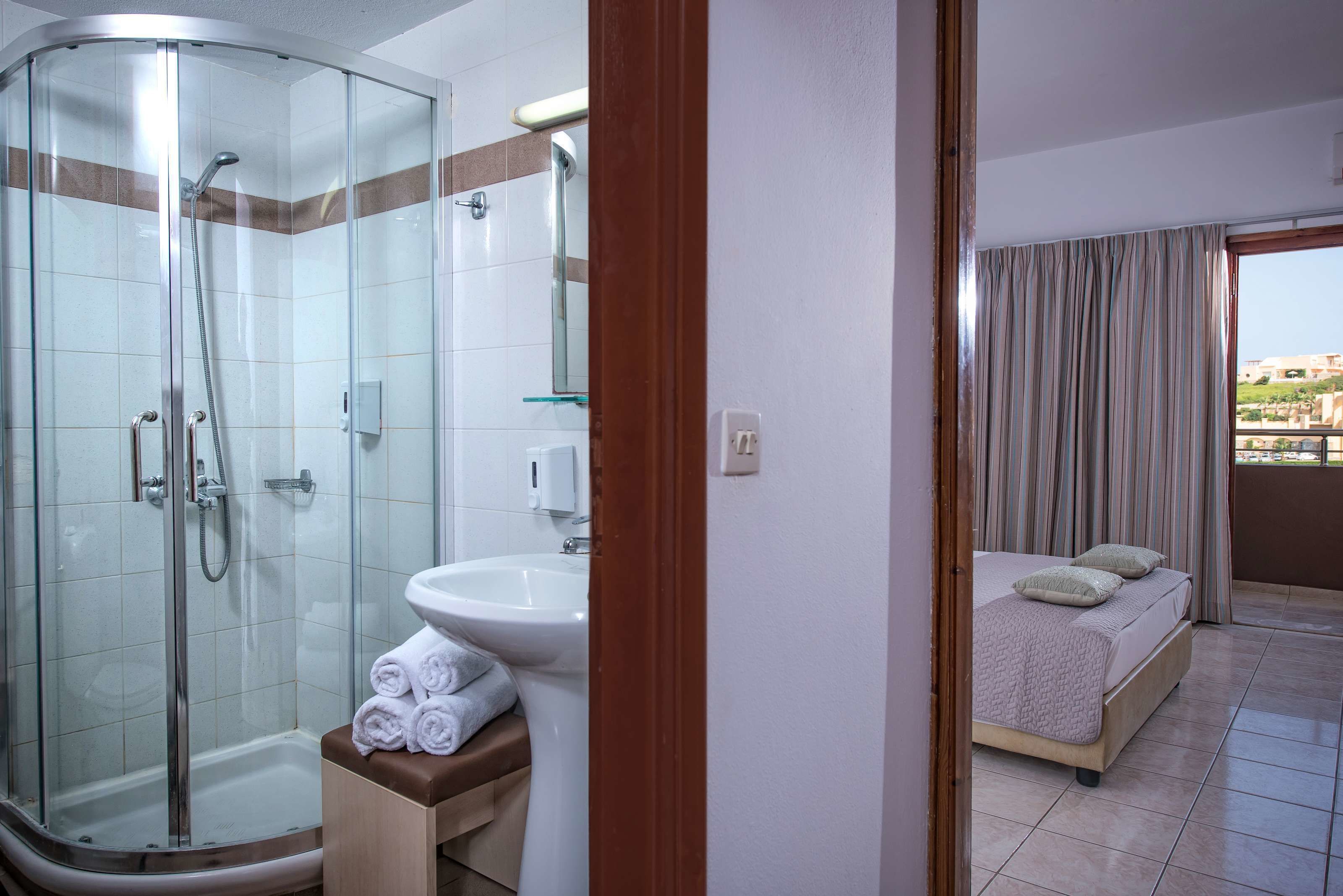 A hotel bathroom with a glass-enclosed shower, a sink, and a bed with a balcony view.