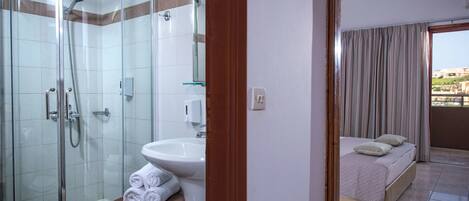 A hotel bathroom with a glass-enclosed shower, a sink, and a bed with a balcony view.