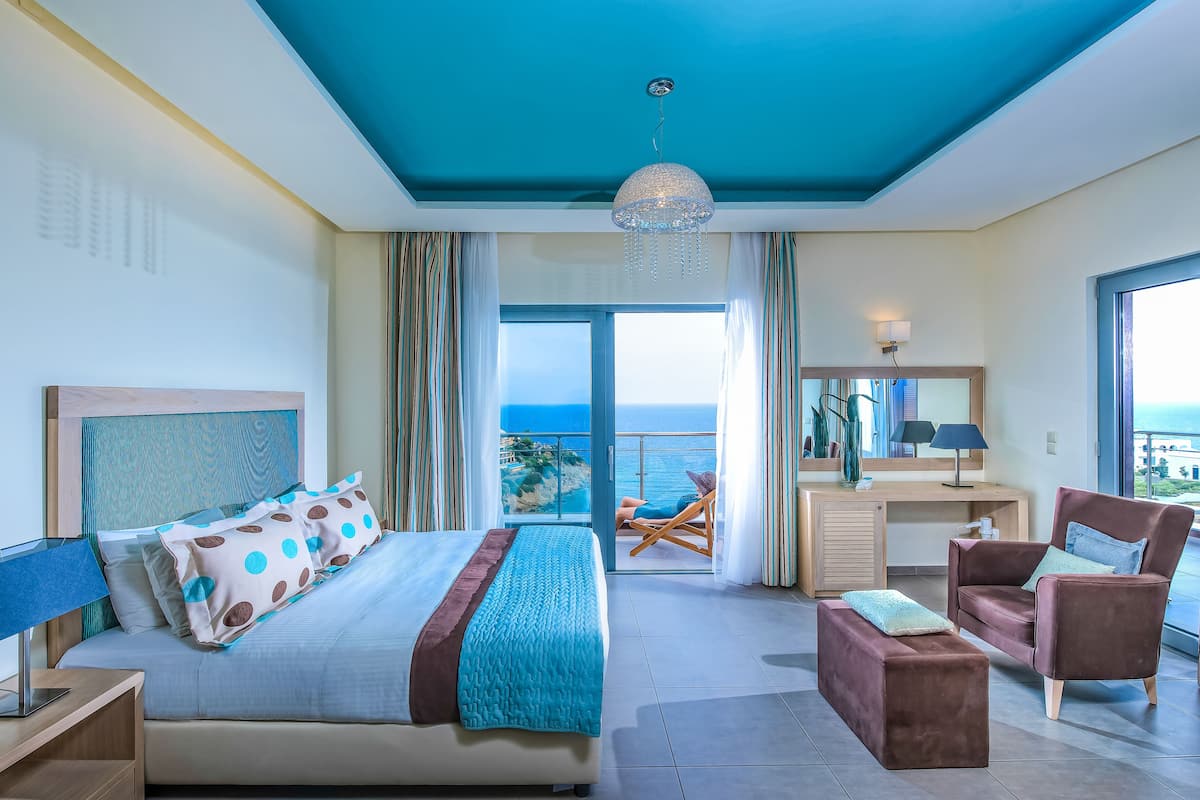 A modern hotel room with a large bed, a desk, a chair, and a balcony with a view of the sea.