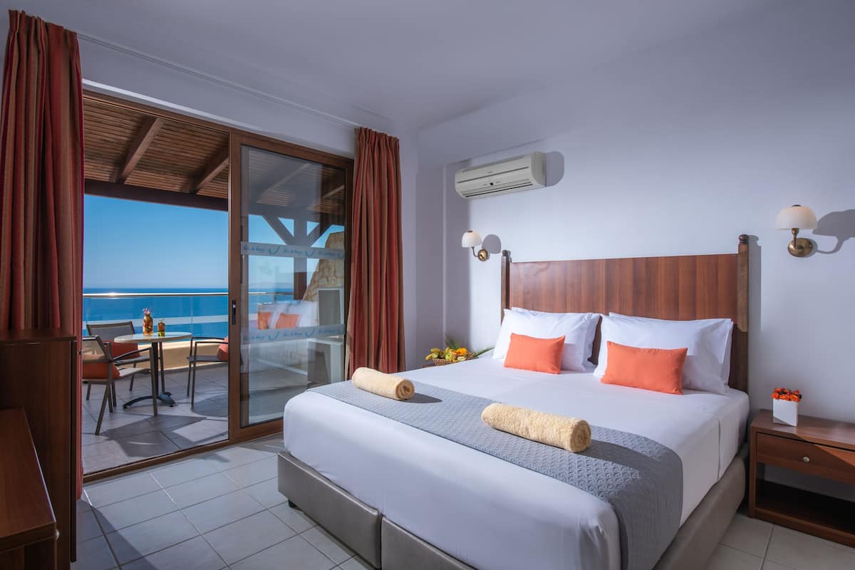 A hotel room with a large bed, a balcony with a view of the sea, a dining area, and an air conditioning unit.