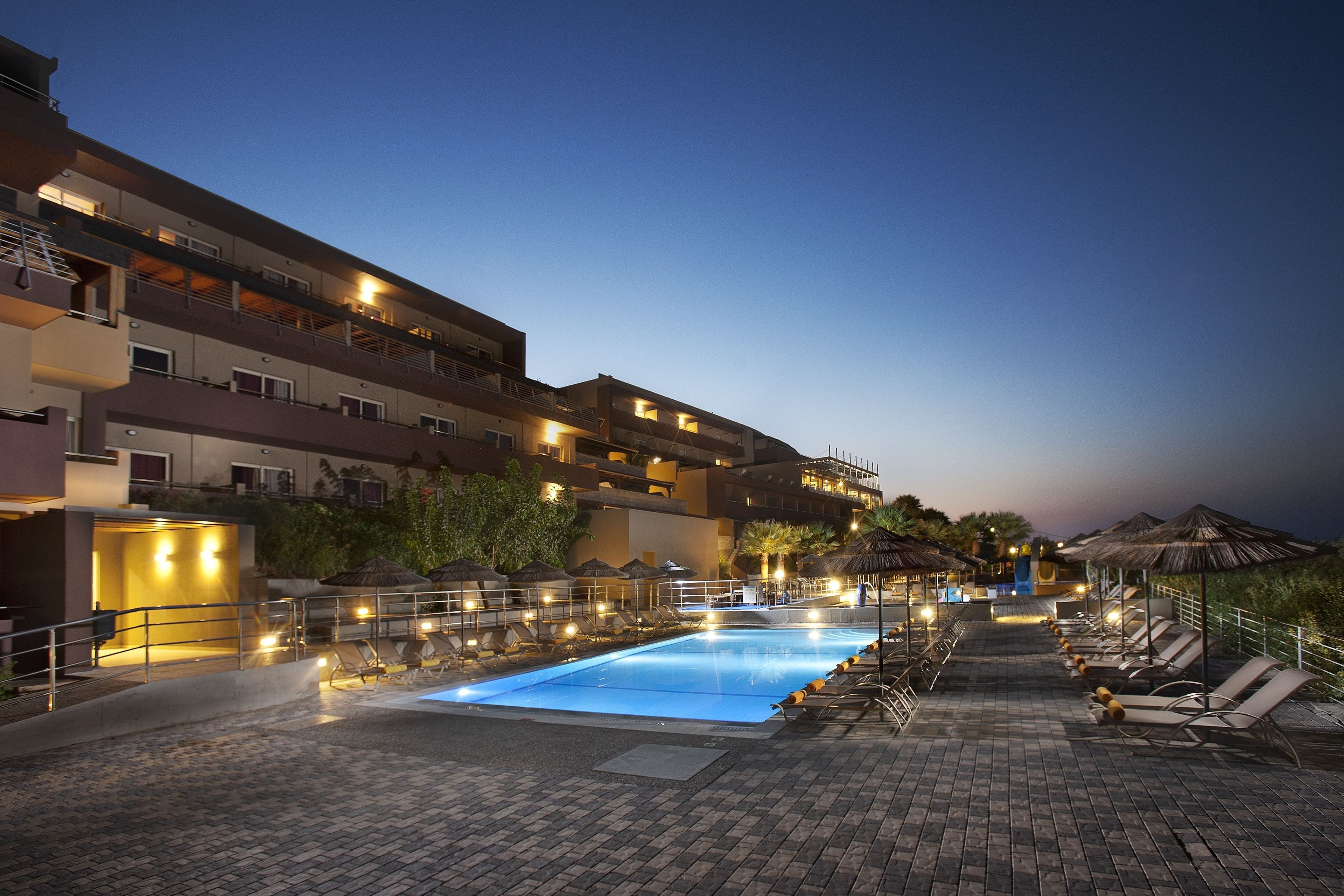 A swimming pool area with lounge chairs and umbrellas, surrounded by a multi-story building with balconies and outdoor lighting.