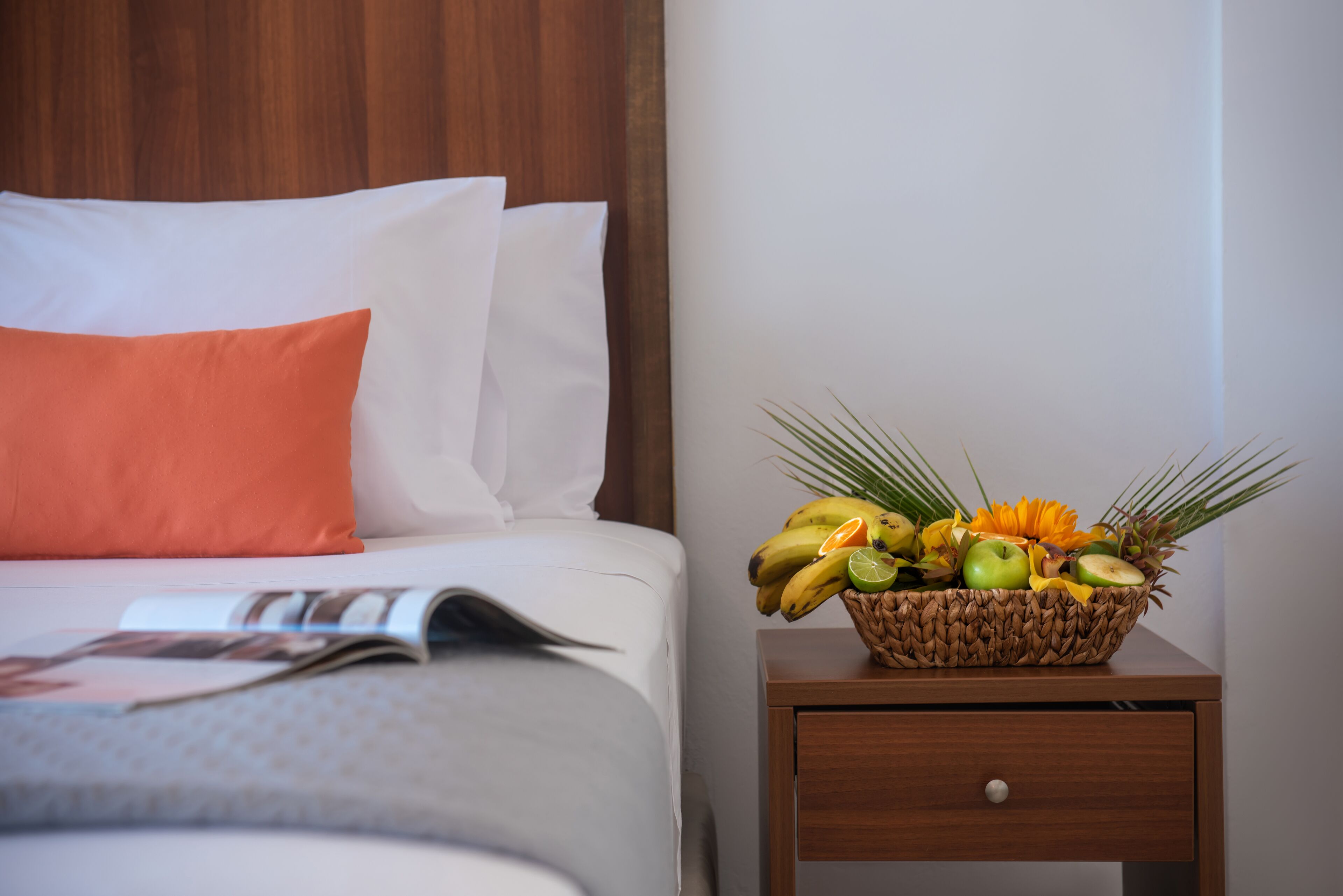 A neatly made bed with white and orange pillows, a magazine, and a fruit basket on a wooden nightstand.