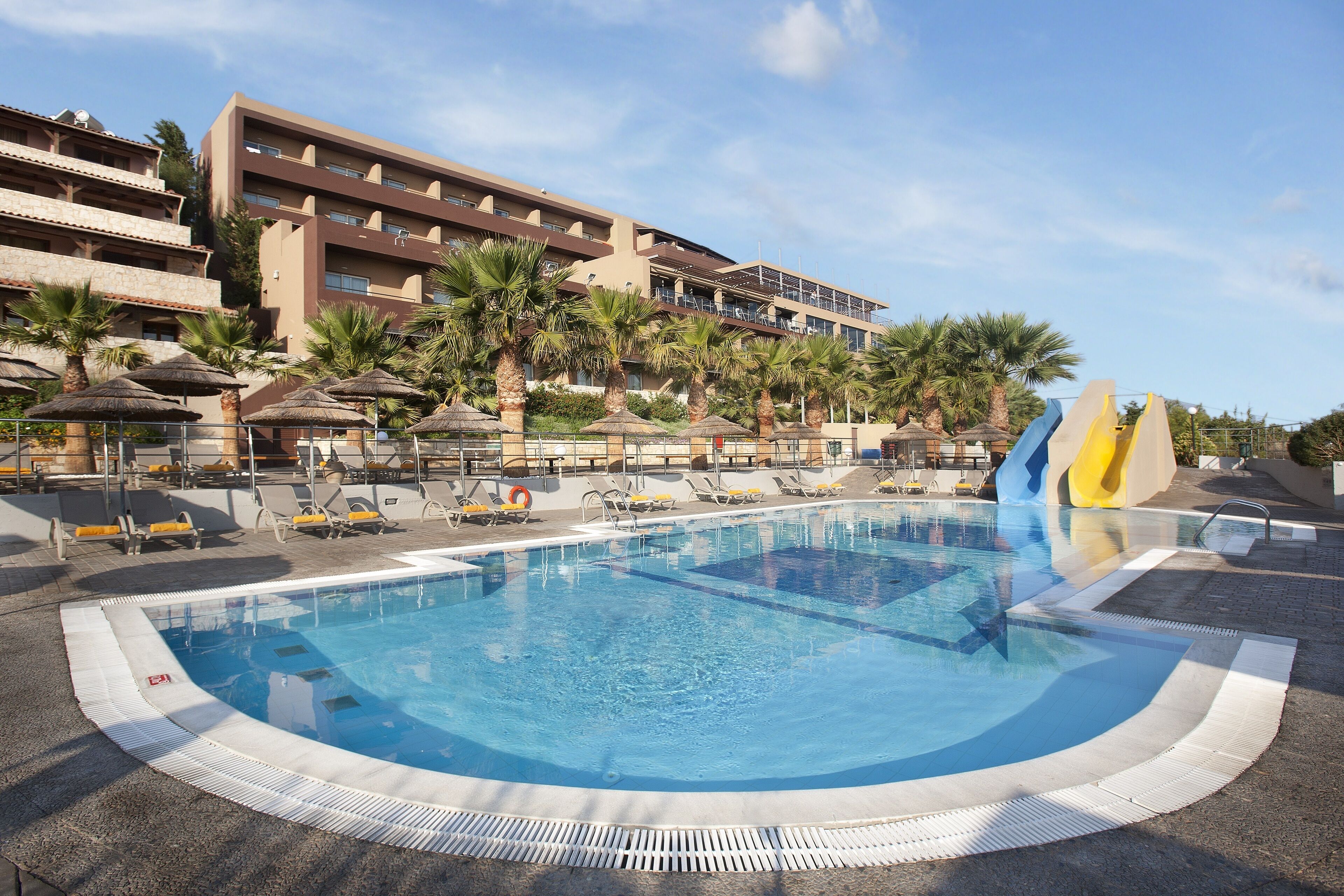 A hotel pool area with a large pool, sun loungers, palm trees, and a water slide.