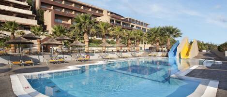 A hotel pool area with a large pool, sun loungers, palm trees, and a water slide.