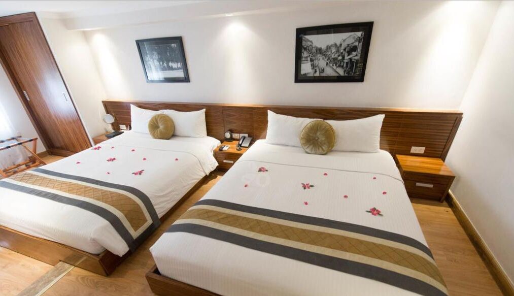 Habitación de hotel con dos camas, cada una con cabecero de madera, ropa de cama blanca y almohada decorativa. Hay cuadros enmarcados en la pared y una puerta de madera a la izquierda.