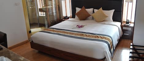 Quarto de hotel bem arrumado com uma cama grande, piso de madeira, box de vidro para o chuveiro e mesa de cabeceira com abajur.