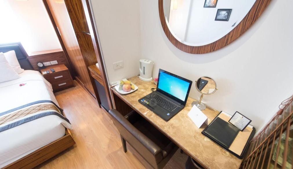 Una habitación de hotel con cama, escritorio con computadora portátil, mesita de noche con lámpara y espejo en la pared.