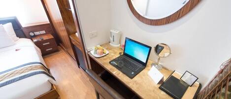 Quarto de hotel com cama, escrivaninha com laptop, mesa de cabeceira com abajur e espelho na parede.