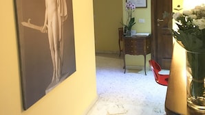 A hallway with a marble floor, a framed painting of a classical sculpture, a wooden door, a small table with a vase of flowers, and a clock on the wall. - Perseo B&B (Rome)