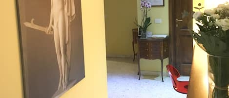A hallway with a marble floor, a framed painting of a classical sculpture, a wooden door, a small table with a vase of flowers, and a clock on the wall.