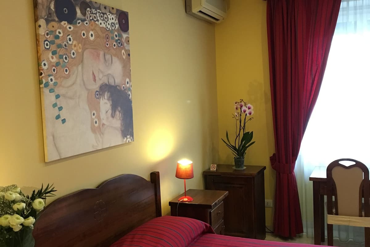 A hotel room with a bed, a nightstand, a desk, a chair, an air conditioner, and a painting on the wall.