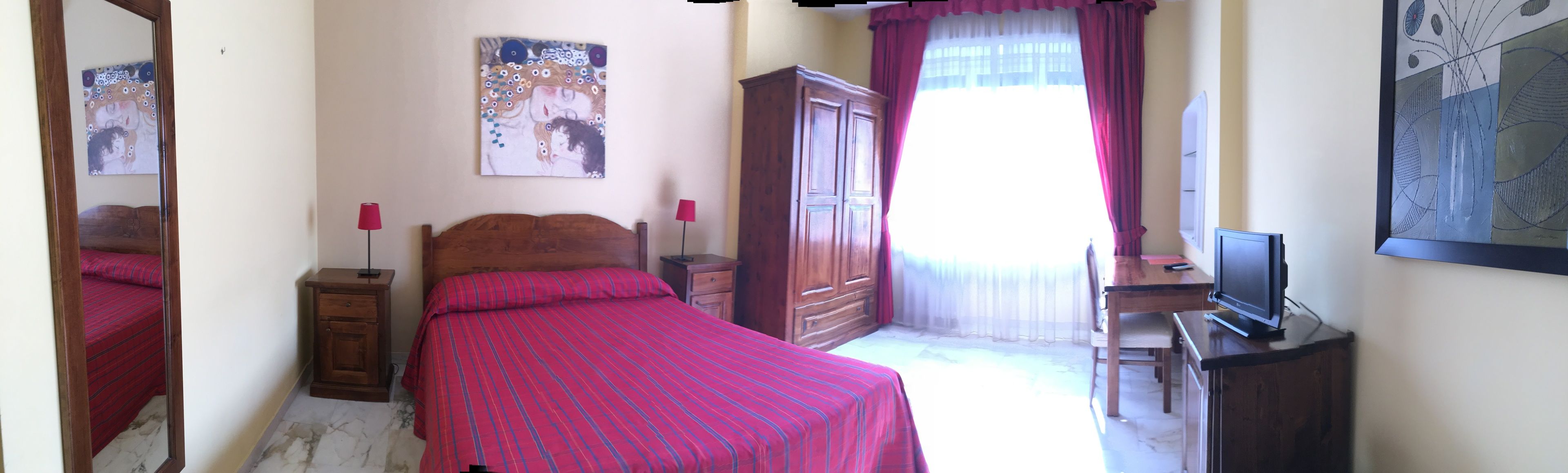 Ein Schlafzimmer mit Bett, Schrank, Nachttisch, Schreibtisch mit Fernseher und Kunstwerken an den Wänden.