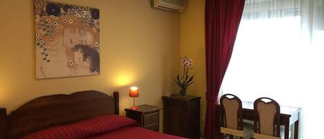 A hotel room with a bed, a desk, chairs, a lamp, an air conditioner, and a painting on the wall.