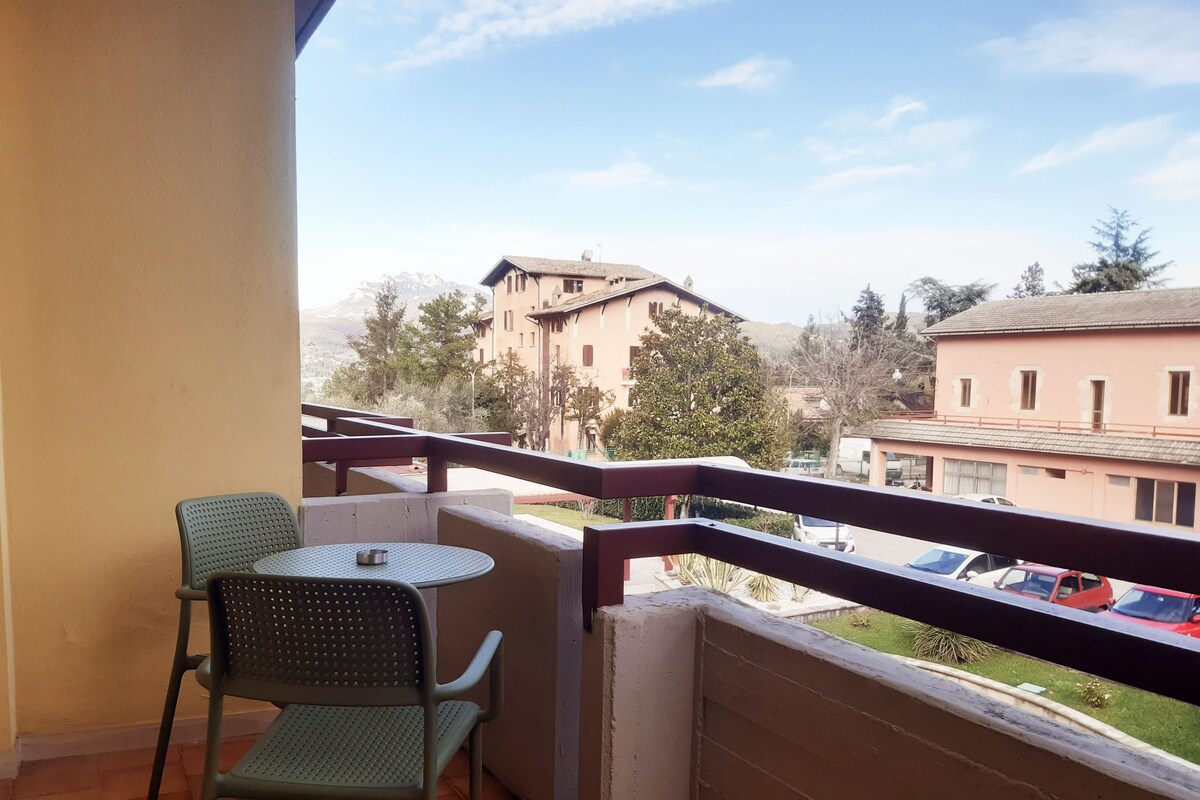 A balcony with a table and two chairs, overlooking a street with parked cars and buildings.
