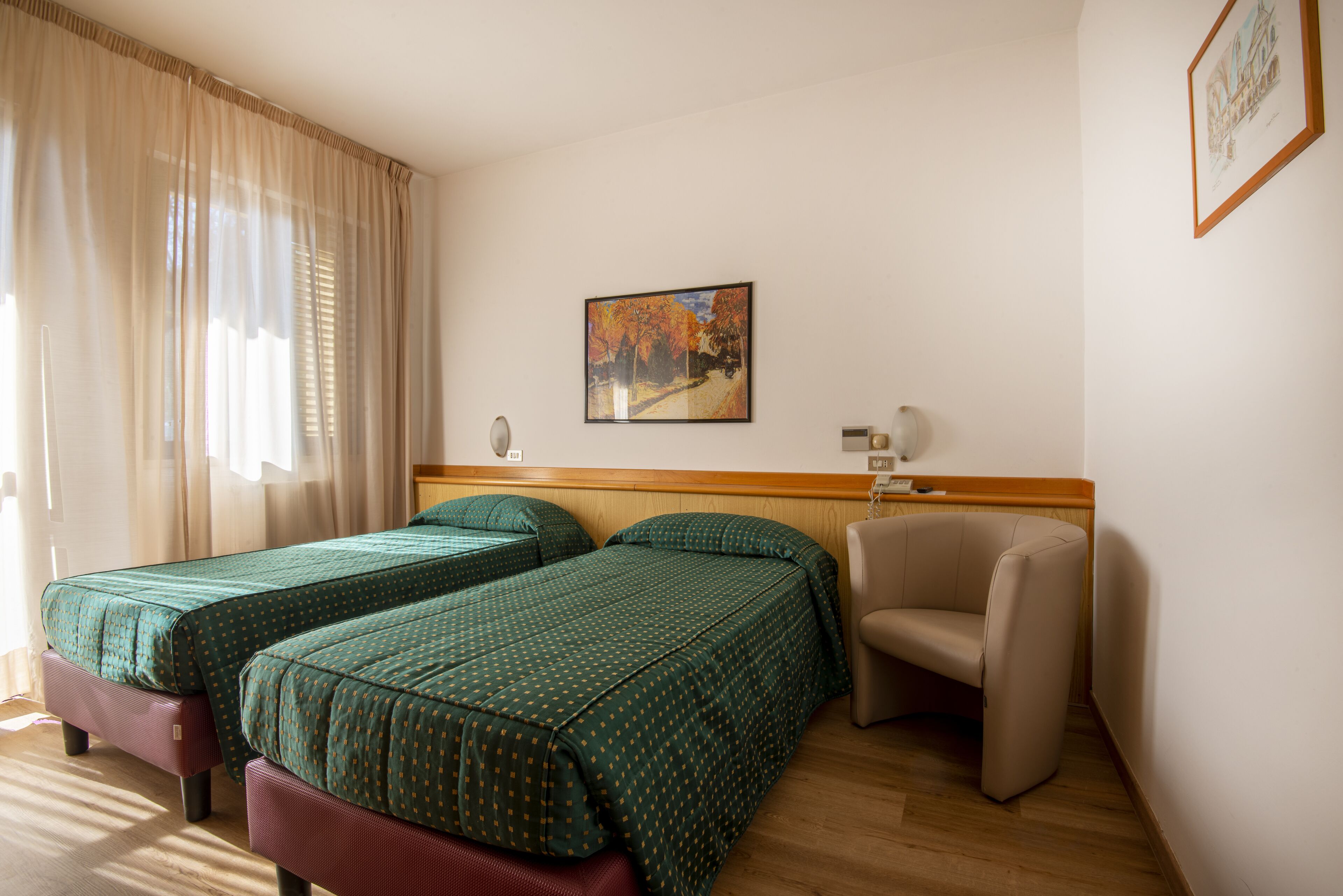 Quarto de hotel com duas camas, uma cadeira, um quadro na parede e uma janela com cortinas.
