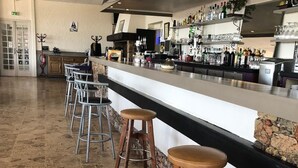 Uma área de bar com piso de ladrilho de pedra, um balcão comprido com banquetas e uma variedade de garrafas e copos.