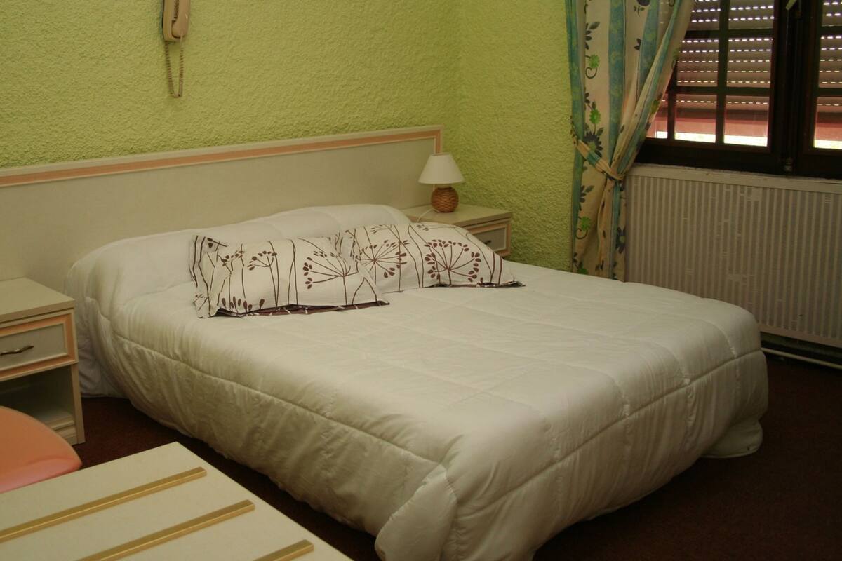 A bedroom with a bed, a nightstand, a lamp, a telephone, and a window with curtains.