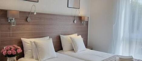 A hotel room with a large bed, bedside tables, a nightstand, a wall-mounted lamp, and a window with sheer curtains.