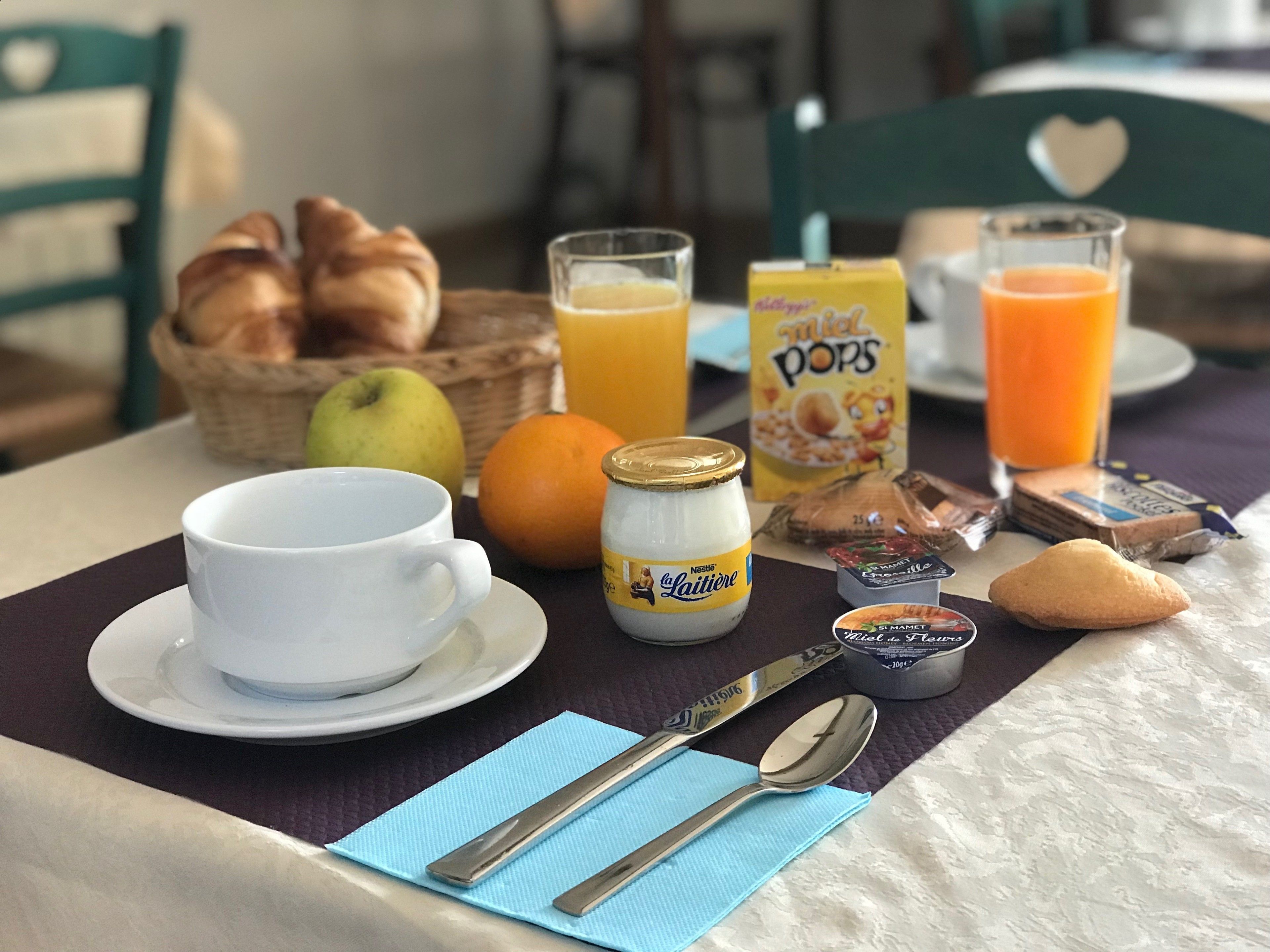 A breakfast table with a cup, a jar of yogurt, a basket of croissants, a glass of orange juice, a carton of milk, and a plate of pastries.