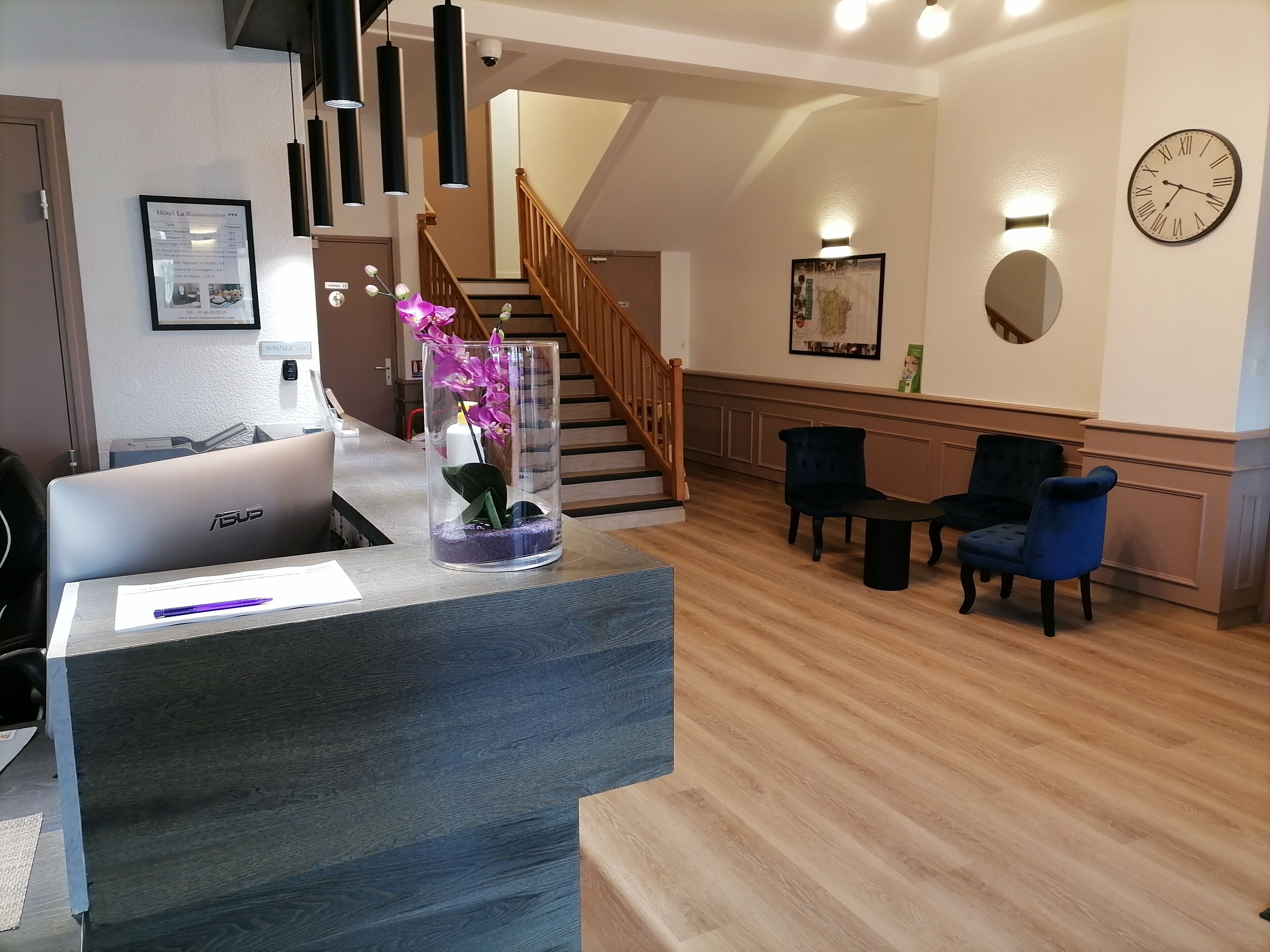 A reception desk with a laptop, a vase with flowers, and a wooden staircase in the background.