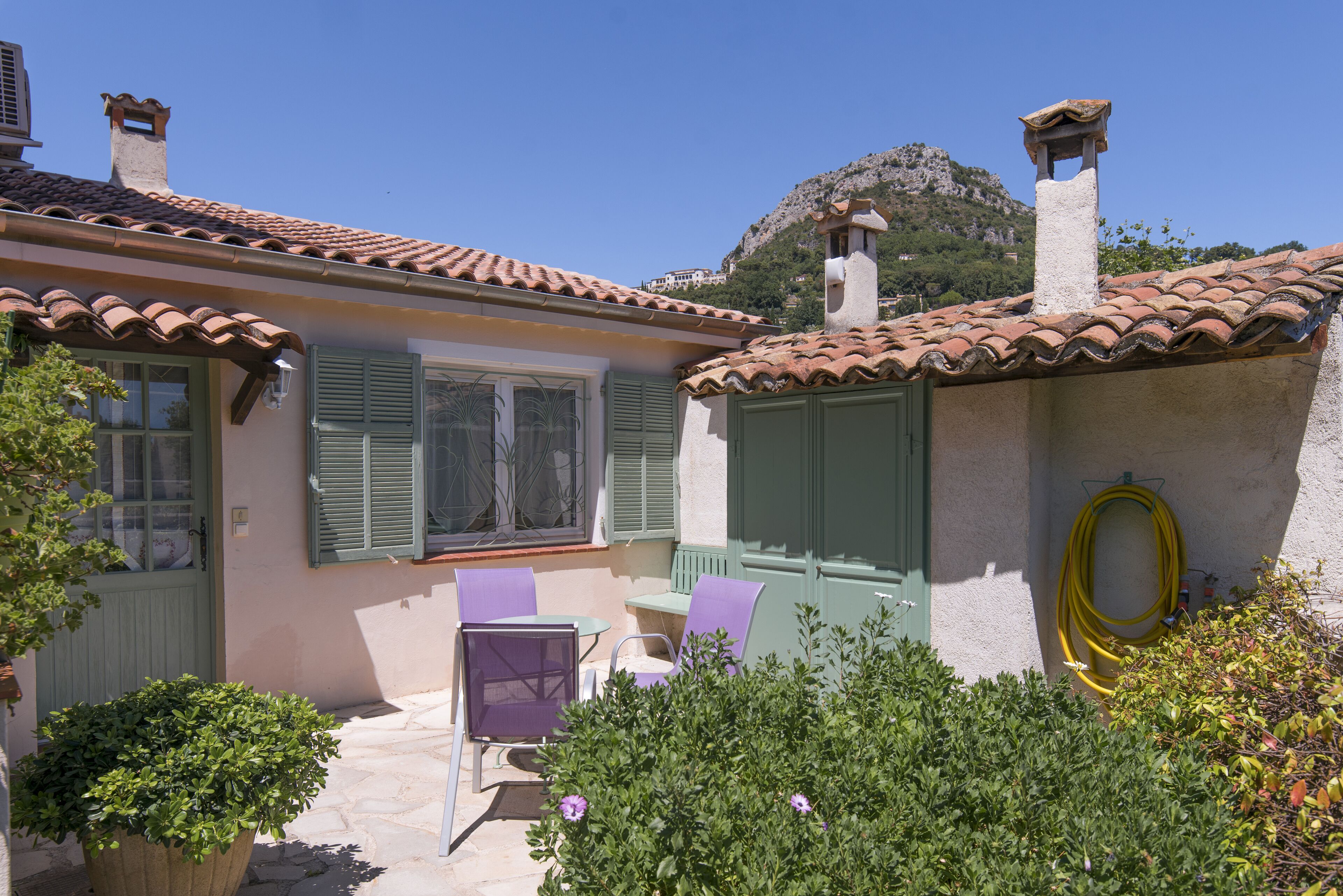 A house with a tiled roof, green shutters, and a garden with a hose.