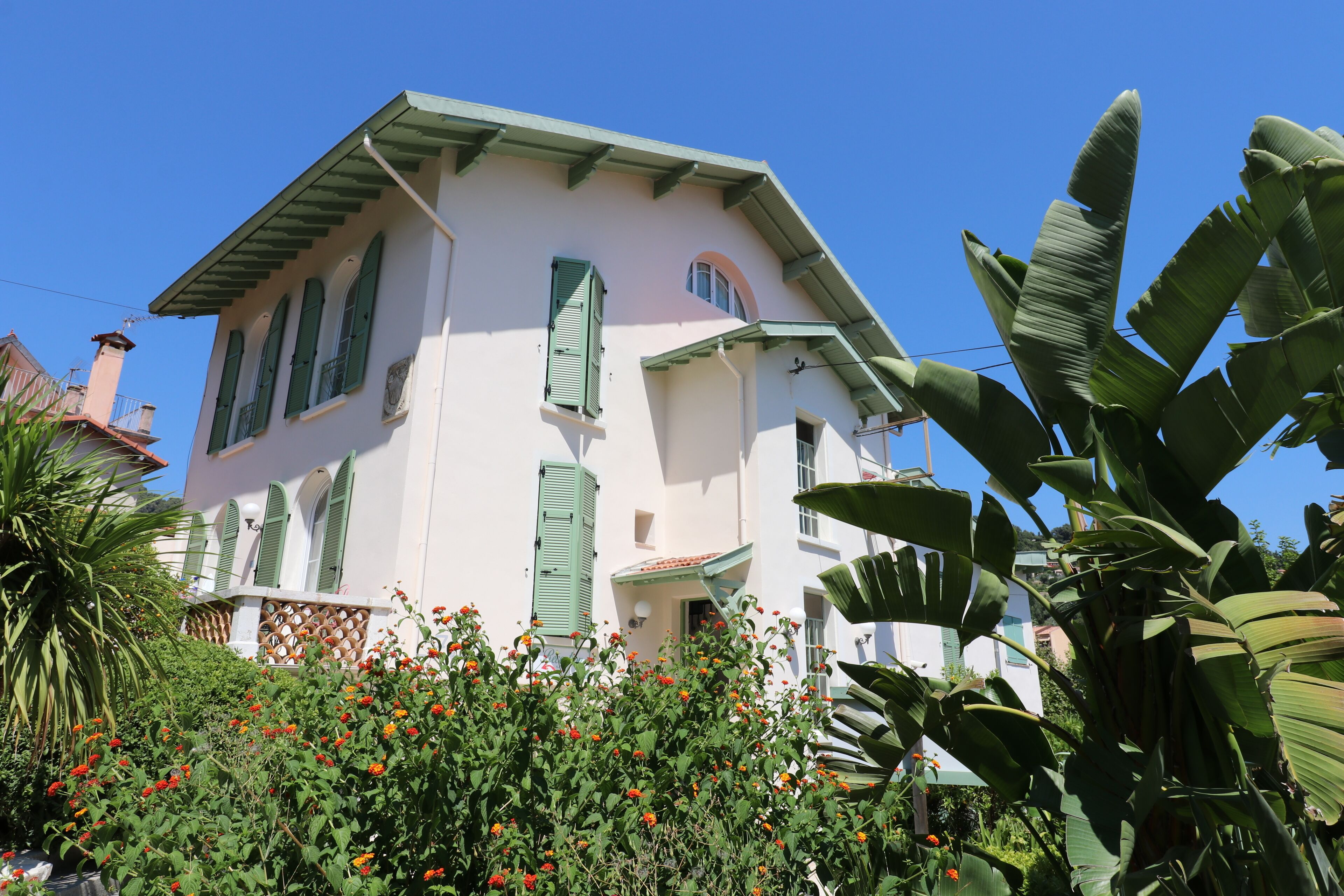 A two-story building with green shutters, a balcony, and a garden with red flowers.