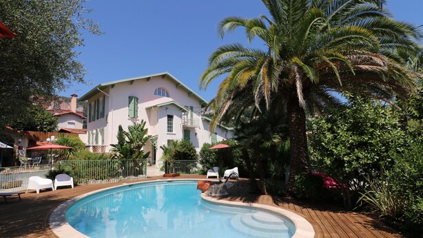 A swimming pool surrounded by a wooden deck, with a house in the background and palm trees. - Hotel Villa Roseraie (Vence)