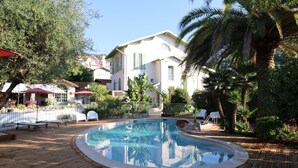 A swimming pool with a wooden deck, surrounded by palm trees and a white building with green shutters. - Hotel Villa Roseraie (Vence)