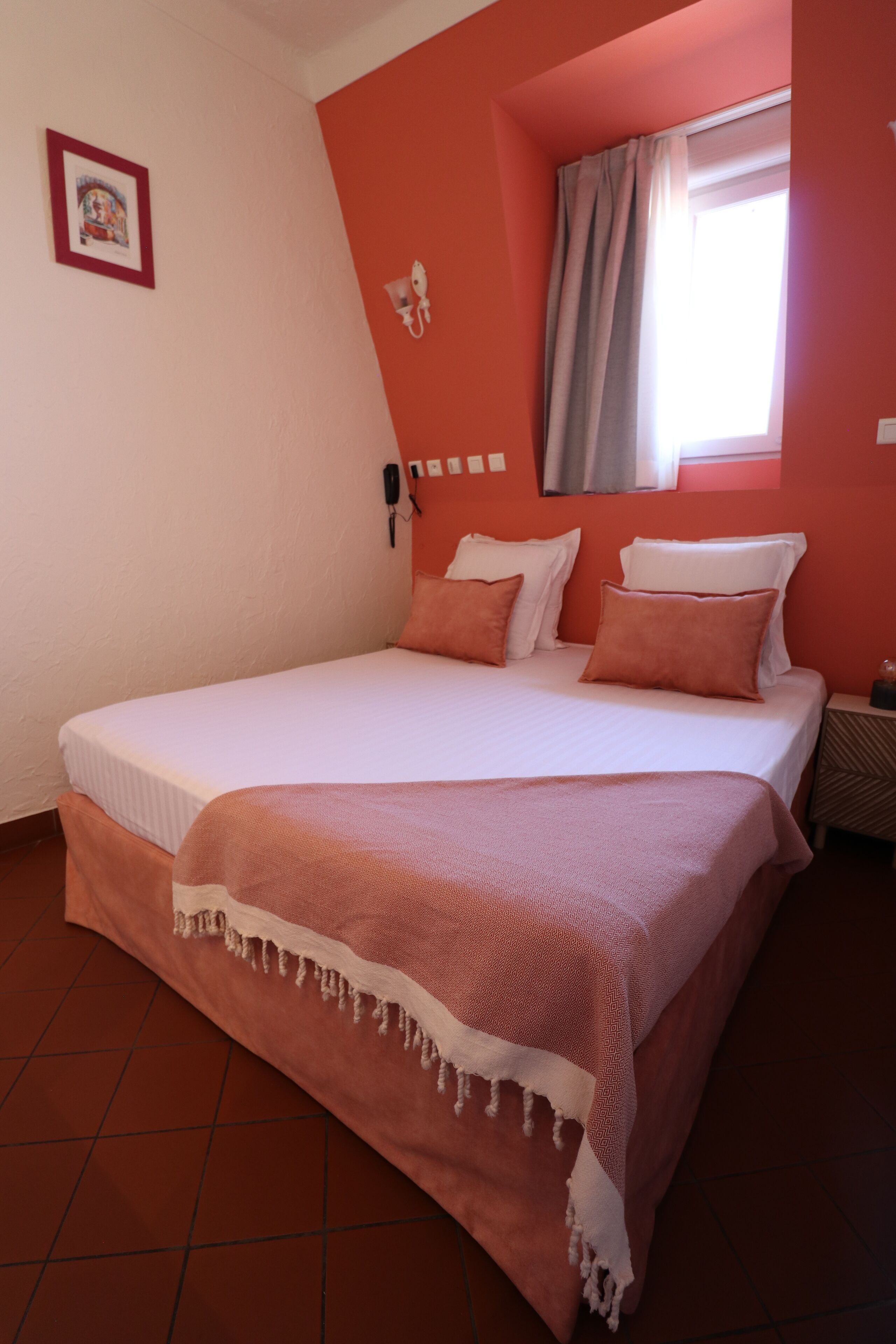 A bed with a white and pink bedspread, a bedside table with a lamp, and a window with curtains.
