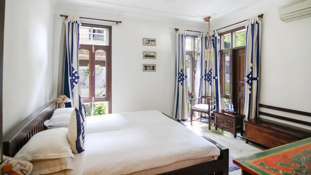 Quarto com cama, escrivaninha e cadeira. Janelas com cortinas, ventilador de teto e ar condicionado na parede.