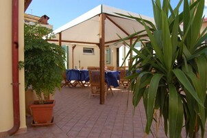 Un patio al aire libre con zona de estar cubierta, plantas en macetas y suelo de baldosas.