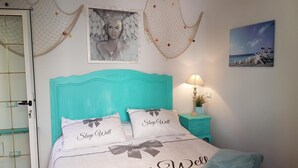 A bedroom with a turquoise headboard, two pillows with 'Sleep Well' text, a framed picture above the bed, a nightstand with a lamp, and a beach scene painting on the wall. - La Papaya (Pisa)